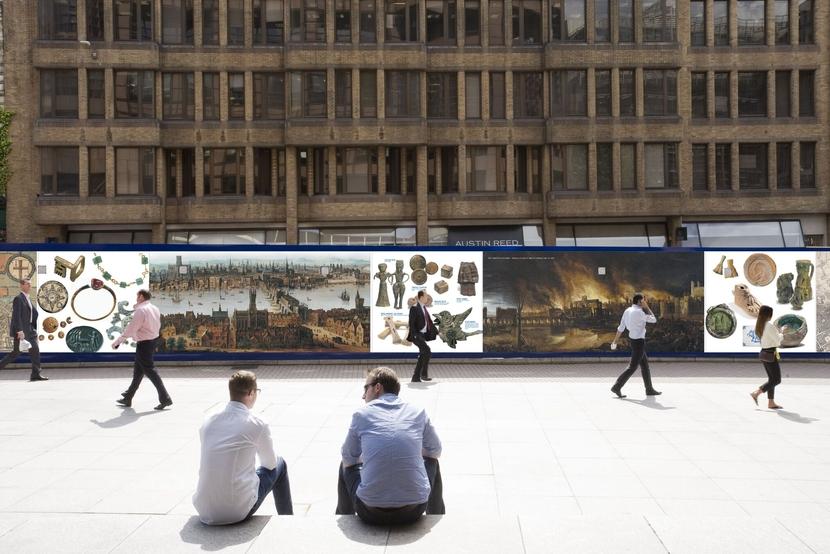 two people sitting opposite a hoarding covered in pictures of archaeological artefacts 