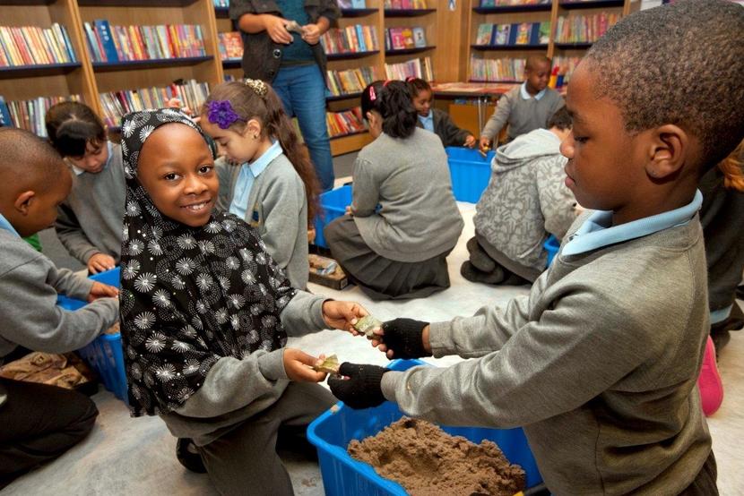 Two primary school children excavate pottery from a sandbox. Other children are in the background engaging in similar activities.