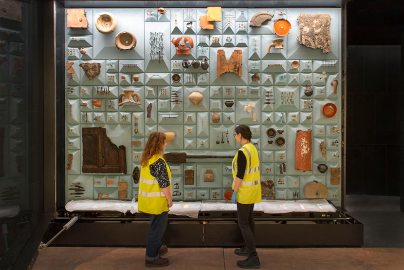 two mola archaeologists standing in front of a large display cabinet containing multiple roman objects.