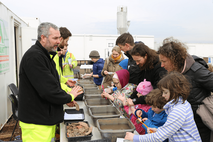 A group of adults and children wash finds using toothbrushes, whilst an archaeologists speak to them.
