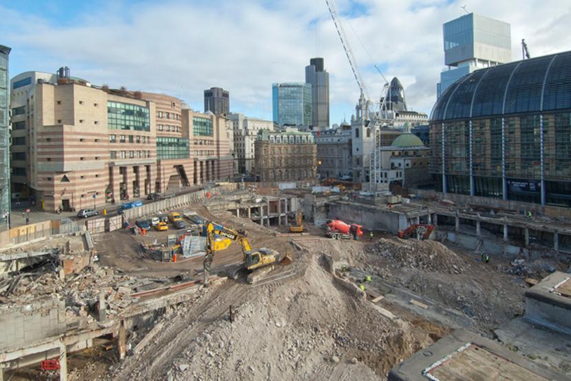 A wide shot of a construction site in the heart of the city of London. There are various bits of machinery on site and a crane in the background.