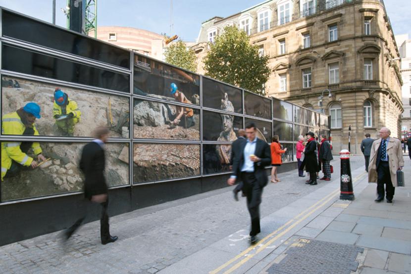 Commuters walk past a set of site hoardings featuring images of archaeological works