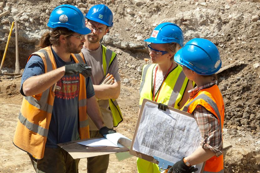 A group of people on a construction site wearing hard hats and hi-vis, in the middle of a discussion. They are holding various archaeological plans.