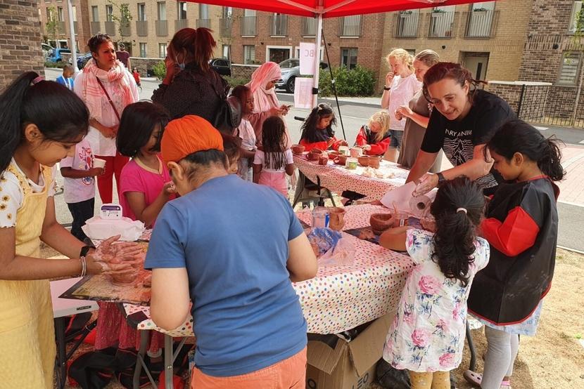 A group of children and adults crowd around a table in the centre of a housing estate. They are making various types of pottery.