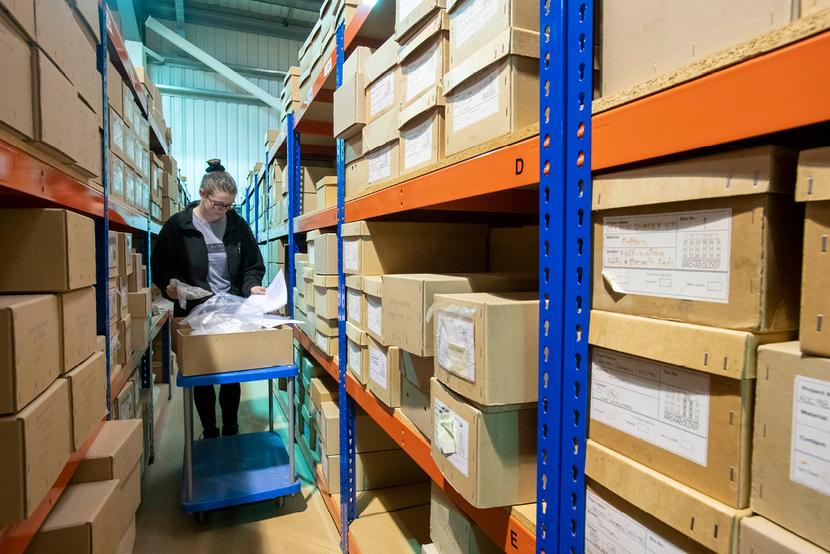 An archaeologist standing in the middle of archive shelves, pushing a small trolley.