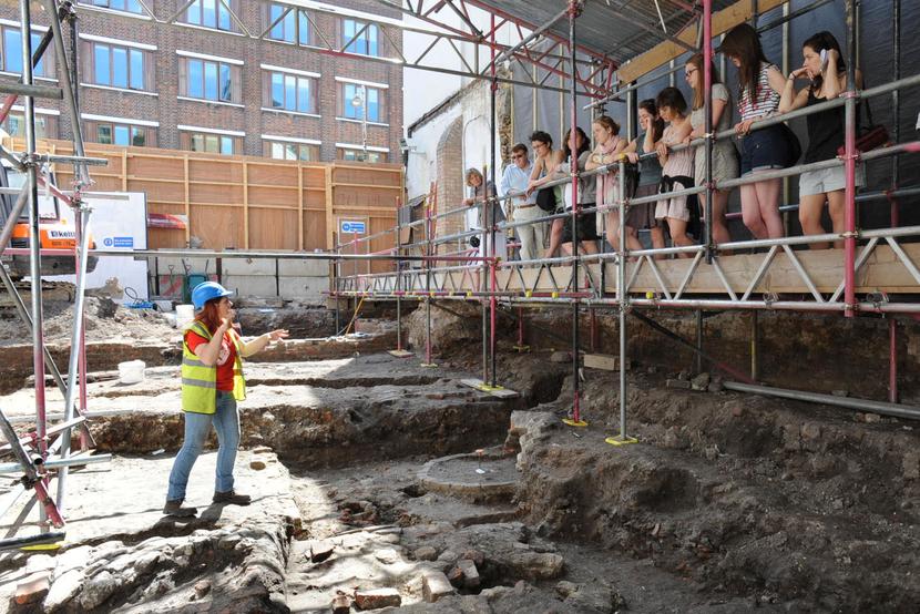 an archaeologist giving a talk on an archaeological site, whilst a group of people listen from a raised viewing platform.
