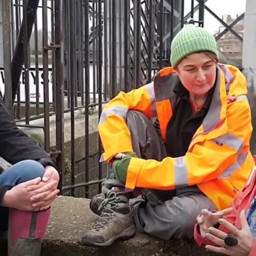 Three women speaking together, they are dressed for the outdoors and are sat on a wall by the River Thames