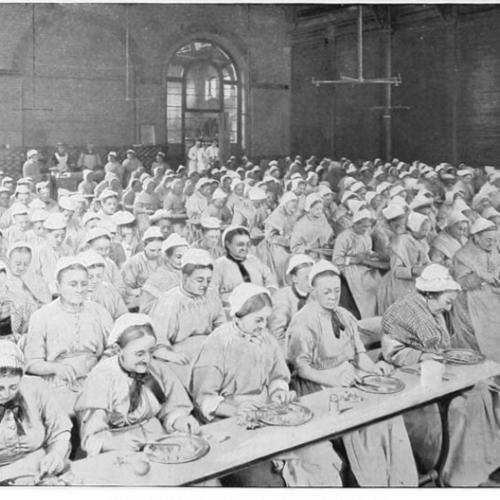 Women wearing white bonnets and victorian clothing eating together in a large dining room. They are seating in rows.